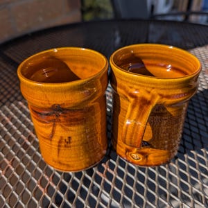 Pode incluir: Duas canecas cilíndricas de cerâmica com um esmalte brilhante cor de mel, apresentando estrias castanhas escuras e uma borda arredondada e lisa. Uma caneca exibe uma alça proeminente no lado direito, enquanto a outra mostra uma superfície lisa.
