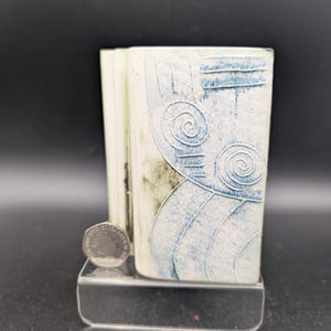 May include: A rectangular, off-white ceramic vase with blue abstract designs, including spirals and lines. The vase is displayed on a clear acrylic stand. A coin rests on the stand. The vase has a textured surface.