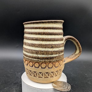May include: A brown and white striped ceramic mug with a decorative pattern around the bottom. The mug has a handle and is sitting on a white surface.