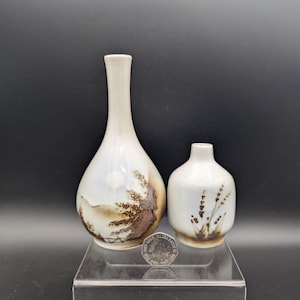 May include: Two ceramic vases. The taller vase is pear-shaped with a long neck, featuring a landscape design in brown and blue. The smaller, round vase has a floral design in brown. Both are on a clear stand.