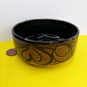 Peut inclure: Un bol en céramique brun avec un intérieur noir et un motif décoratif de volutes et de cercles bruns. Le bol est posé sur une surface jaune.