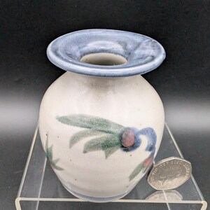 Op de afbeelding: Een kleine, ronde keramische vaas met een brede, blauwe rand. De gebroken witte body is versierd met groene en blauwe bloemmotieven. De vaas wordt gepresenteerd op een transparante acrylstandaard tegen een zwarte achtergrond.