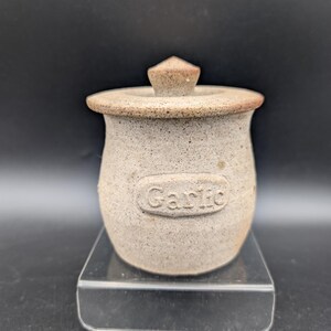 Peut inclure: Un pot à ail en céramique beige moucheté avec un couvercle assorti. Le récipient présente un corps arrondi et une étiquette rectangulaire en relief sur le devant avec le mot Garlic visible en lettres texturées sur la surface en argile.