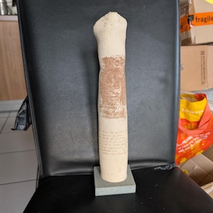 May include: A beige, cylindrical sculpture with a textured, brown lower section and a smooth, off-white upper portion. The sculpture is mounted on a small, grey rectangular base. Faint text is visible on the lower portion of the sculpture.