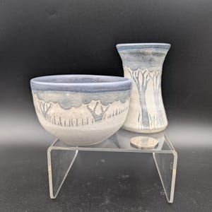 Könnte beinhalten: Ein Keramikschalen- und Vasen-Set. Die Schale ist rund mit blauem Rand und Landschaftsdesign. Die Vase ist hoch mit einem ähnlichen Design. Beide sind weiß mit blauen Akzenten. Das Set wird auf einem transparenten Acrylständer präsentiert.