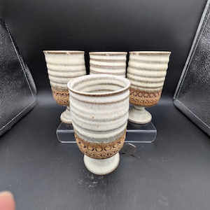 May include: Four ceramic goblets in cream and brown. The goblets have a footed base and a textured, striped design. The lower portion of the goblets features a decorative pattern. The background is black.