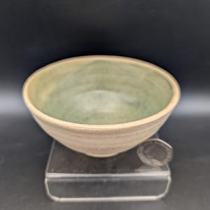 May include: A round ceramic bowl with a light beige exterior and a mottled sage green interior glaze. The bowl features visible horizontal throwing lines along the side and is positioned next to a small circular coin for scale reference.