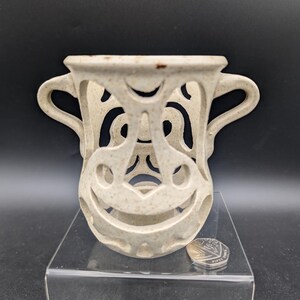 May include: A beige ceramic vase with a unique openwork design. The vase has two handles and a face-like pattern created by cut-out shapes. The vase is displayed on a clear stand, with a coin next to it for scale.