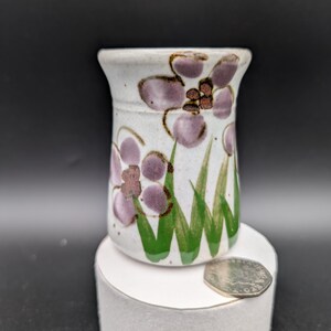 May include: A small white ceramic vase with a floral design. The vase has purple flowers with brown centres and green leaves painted on it.