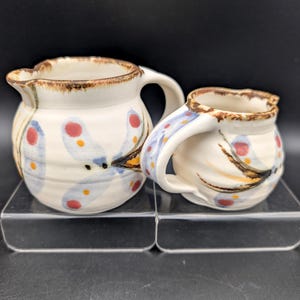 May include: Two cream-coloured ceramic pitchers with a rounded shape and a handle on the side. The pitchers feature a hand-painted pattern of blue, red, and yellow dragonfly-like motifs and a brown, textured rim around the top opening.