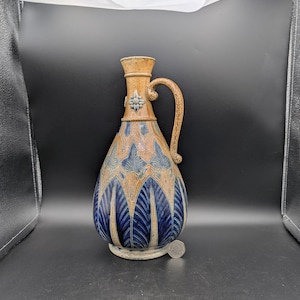 Könnte beinhalten: Ein Keramikkrug mit einem beigefarbenen Griff und Hals sowie einem bauchigen Körper. Der Körper ist mit blauen und beigefarbenen blattartigen Mustern verziert. Der Krug hat einen kleinen, runden Sockel und ein dekoratives Blumendesign in der Nähe des Griffs.