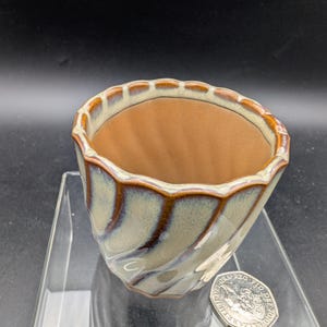 May include: A ceramic plant pot with a distinctive, spiralled design. The exterior has a mix of blue, brown, and cream glazes, with a warm brown interior. The rim has a scalloped edge. A coin is next to the pot.