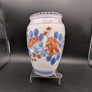 Könnte beinhalten: Eine weiße Keramikvase mit breiter Öffnung, verziert mit blauen und rostfarbenen Blumenmustern. Die Vase hat ein blaues Band am Boden und am Rand und wird auf einem transparenten Ständer ausgestellt. Die Vase ist etwa 18 cm hoch.