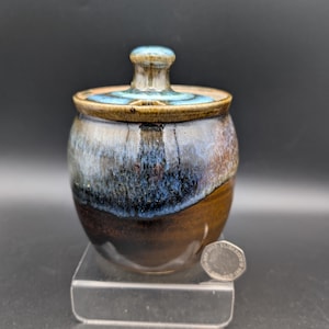 May include: A small, handcrafted ceramic jar with a lid. The jar is a rounded shape, with a brown base that fades into blue and white hues. The lid has a small knob and a blue rim. The jar is displayed on a clear stand.