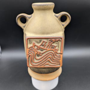 May include: A small, tan ceramic vase with two handles. The vase has a raised, rectangular design on the front depicting a ship sailing on waves.