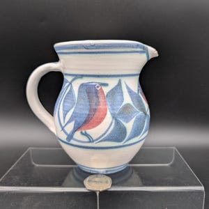 以下が含まれることがあります： 白ベースと取っ手が付いたセラミックピッチャー。青と赤の鳥と葉のデザインで飾られています。ピッチャーには注ぎ口と青いストライプのリムが付いています。手描きのデザインが、ユニークで芸術的な外観を与えています。