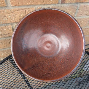 Op de afbeelding: Een grote, ronde keramische kom met een gespikkeld, roodbruin interieur en een donkerdere rand. De kom heeft een glanzende afwerking en staat op een zwart metalen oppervlak. De buitenkant is niet zichtbaar.