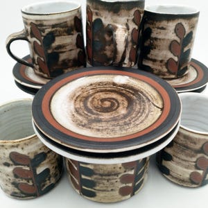 May include: Set of four brown and white ceramic coffee mugs with a stylised leaf design. The mugs are stacked on top of each other with matching saucers.