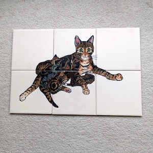 May include: A set of four white ceramic tiles with a hand-painted illustration of two brown tabby cats. The larger cat is sitting with its front paws outstretched, while the smaller cat is lying down with its head resting on the larger cat's back.
