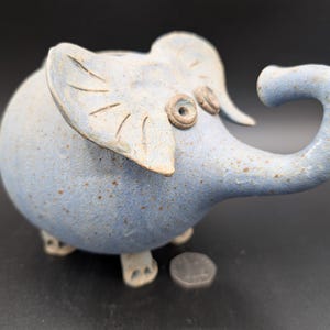 May include: A light blue ceramic elephant figurine with a curled trunk and large ears. The elephant has a round body and small feet. The figurine is speckled with brown spots and has a coin slot on top. A coin is next to the elephant.