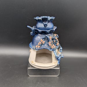 Puede incluir: Un calentador de aceite de cerámica con una sección superior esmaltada en azul profundo con forma de flor, sobre una base blanca con una abertura arqueada. La sección azul está decorada con pequeños capullos de rosa rosados y enredaderas.