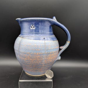 May include: A ceramic jug with a blue glaze and a textured, light blue and orange-brown body. The jug features a decorative band of small indentations near the rim, a three-circle stamp, and a curved handle. A silver coin is placed at the base.
