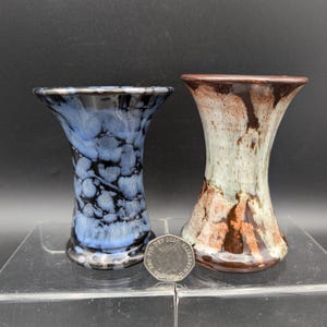 Könnte beinhalten: Zwei kleine Keramikvasen mit ausgestelltem Rand und Sanduhrform. Eine Vase hat eine blau-schwarze gesprenkelte Glasur, die andere eine cremefarbene und braune Tropfglasur. Eine britische Zwanzig-Pence-Münze liegt zum Größenvergleich dazwischen.