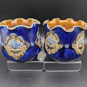 May include: Two matching blue ceramic planters with a ruffled rim and ornate floral designs. The planters have a dark blue glaze with brown and gold accents. Each planter features a central floral motif.