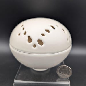 Puede incluir: Una esfera de cerámica blanca con una serie de agujeros irregulares, posiblemente un objeto decorativo o un difusor. La esfera se encuentra sobre un soporte transparente, con una moneda visible para la escala. El objeto tiene un acabado liso y mate.