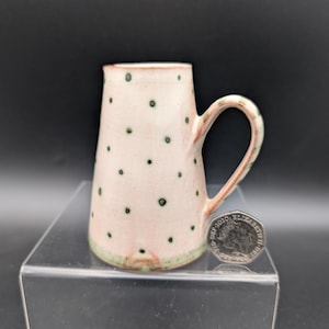 May include: A handmade ceramic pitcher with a pinkish-white body and small, dark green polka dots. The handle and rim have a reddish-brown hue, and the base is trimmed with green. A coin is next to the pitcher.
