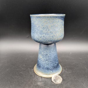 Op de afbeelding: Een blauwe keramische kelk met een gespikkelde glazuur. De kelk heeft een brede, ronde kom en een hoge, slanke steel.