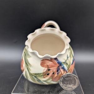 May include: A small, off-white ceramic bowl with a ruffled rim and a handle. The bowl is decorated with orange and blue floral designs and green leaves. A coin is next to the bowl.