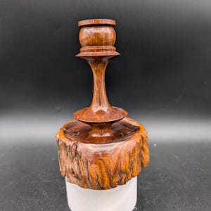 May include: A wooden candle holder with a natural wood finish. The holder has a round base and a tall, slender stem. The top of the holder is a round, open cup for holding a candle.