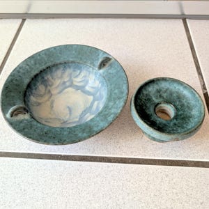 May include: Two-piece ceramic ashtray set. The ashtray is round with a teal exterior and a light blue and white interior. The set includes a matching lid with a central hole. The ashtray has two small grooves for resting cigarettes.
