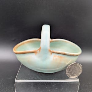 May include: A small ceramic basket with a light blue and green glaze, featuring a single arched handle and a brown rim. The basket is positioned next to a British twenty pence coin for scale, which provides a clear visual reference for its size.