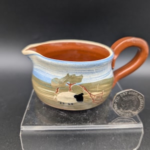 May include: A small, ceramic creamer with a brown handle and rim. The body features a landscape design with a sheep and tree. The creamer is approximately 3 inches in diameter and 2 inches tall.