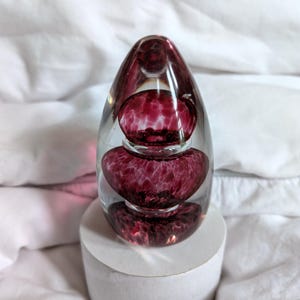 May include: A clear glass paperweight with three internal, layered orbs of deep red, speckled glass. The paperweight is egg-shaped and sits on a white cylindrical base. The glass has a glossy finish.
