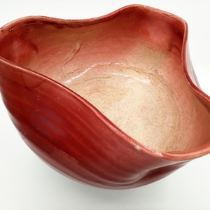 Può includere: Una ciotola in ceramica rossa con una forma unica, ondulata e organica. La ciotola ha una finitura liscia e lucida e un interno beige chiaro.