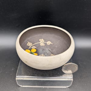 May include: A shallow, round ceramic bowl with a matte off-white exterior and a dark brown interior. The interior features a hand-painted design of a yellow flower with green leaves on a thin vine. A silver coin is placed next to the bowl for scale.