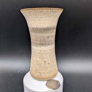 May include: A handcrafted ceramic vase with a speckled beige exterior and a flared top. The vase features bands of grey and brown hues, adding a rustic touch. The vase is approximately 15 cm tall.