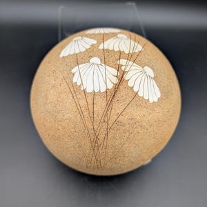 May include: A round, tan ceramic piece with a textured surface. White daisy-like flowers with thin stems are etched into the surface. The piece is displayed against a dark background.
