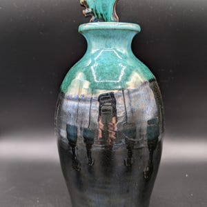 Könnte beinhalten: Eine handgefertigte Keramikflasche mit türkisfarbener und schwarzer Glasur. Die Flasche hat einen dekorativen türkisfarbenen Verschluss mit einem einzigartigen, strukturierten Design. Der Flaschenkörper geht von Schwarz zu Türkis über und zeigt die Fähigkeit des Kunsthandwerkers.