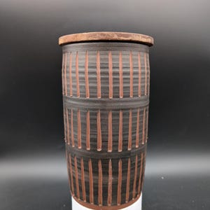 May include: A cylindrical ceramic vase with a brown and black striped pattern. The vase has a white base and a lid.