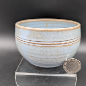 May include: A light blue ceramic bowl with a speckled finish and a brown rim. The bowl features horizontal ridges along the exterior. A British twenty pence coin is placed next to the bowl for scale, showing the relative size of the ceramic vessel.