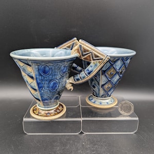 Op de afbeelding: Twee keramische mokken met blauw glazuur en geometrische patronen, waaronder cirkels en ruiten. De mokken hebben hoekige, getextureerde handvatten en uitlopende randen, rustend op ronde voetstukken. Een munt is zichtbaar voor de schaal.