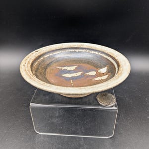 Könnte beinhalten: Eine kleine, weiße Keramikschale mit brauner Glasur und einem weißen Design aus zwei Vögeln. Die Schale steht auf einem klaren Acryl-Ständer.