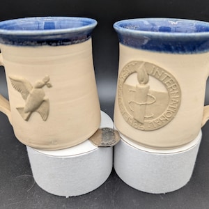 May include: Two ceramic mugs with blue rims. One mug features a dove with an olive branch, the other has a raised logo that reads "University of Notre Dame 1842 International".