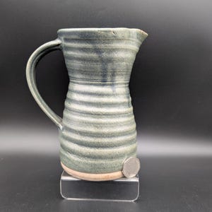 Puede incluir: Jarra de cerámica hecha a mano con asa curva y boquilla. La jarra es de color azul verdoso apagado con detalles horizontales acanalados. La base es de color marrón claro. La jarra se exhibe sobre un soporte transparente.