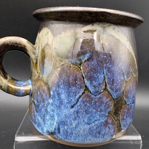 May include: A ceramic mug featuring a crystalline glaze with deep blue, indigo, and earthy brown tones. The surface displays a distinct pattern of large, circular crystal formations with dark edges, a wide handle, and a dark, textured rim.