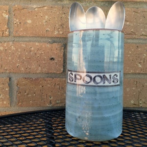 May include: A blue ceramic spoon holder with the word "SPOONS" written on the front. The holder is filled with silver spoons.